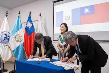 Dos hombres firman documentos sobre una mesa azul, una mujer observa. Al fondo, banderas de Guatemala, Taiwán y del Ministerio de Salud. Proyección de banderas