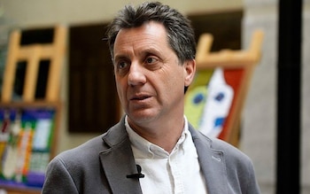 Primer plano de un hombre de mediana edad, con cabello oscuro, saco gris, camisa blanca y micrófono de solapa. Al fondo, objetos de madera y pinturas abstractas