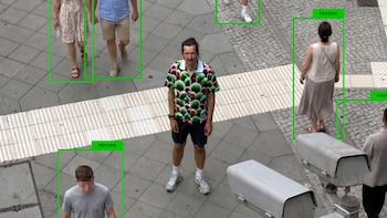 Vista aérea de personas caminando en una calle peatonal con recuadros verdes de detección digital y una cámara de vigilancia en primer plano