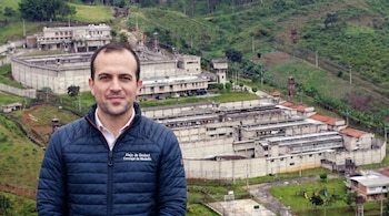 Retrato de Alejo de Bedout, con una chaqueta azul, posando frente a una prisión con altos muros de hormigón y torres de vigilancia en un entorno montañoso.