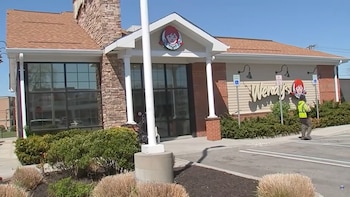 Vista exterior de un restaurante Wendy's con techo marrón, fachada de ladrillo y paredes beige. Hay arbustos en primer plano y una persona caminando