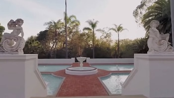Vista de una piscina ornamental con una fuente central y caminos de ladrillo, flanqueada por dos estatuas de estilo clásico sobre pedestales blancos