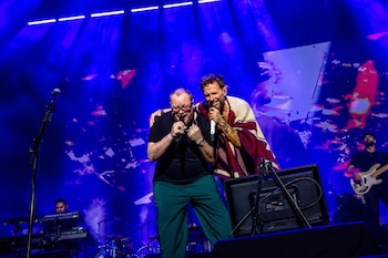 Dos hombres de Sin Bandera cantan juntos en un escenario iluminado de azul. Uno usa un poncho, el otro camiseta negra, con micrófonos y pantallas de fondo
