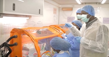 Cuatro personas con ropa de protección y mascarillas manipulan objetos en cabinas de aislamiento de color naranja