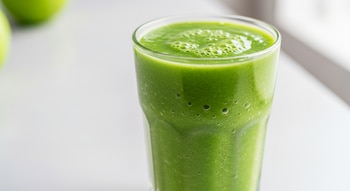 Un vaso transparente con licuado verde cremoso de manzana y apio, con pequeñas burbujas en la superficie. El vaso está sobre un fondo blanco y minimalista.
