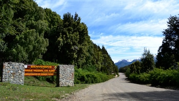 Cartel de madera del Parque Nacional Los Alerces junto a un camino de tierra bordeado por árboles y montañas con nieve de fondo