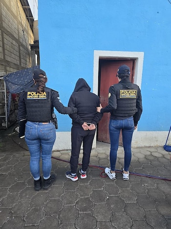 Vista trasera de dos agentes de policía con chalecos tácticos escoltando a una persona esposada vestida de negro, frente a una pared azul