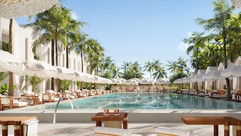 Piscina exterior de un hotel con agua cristalina, flanqueada por hileras de tumbonas, sombrillas blancas y palmeras altas, bajo un cielo azul