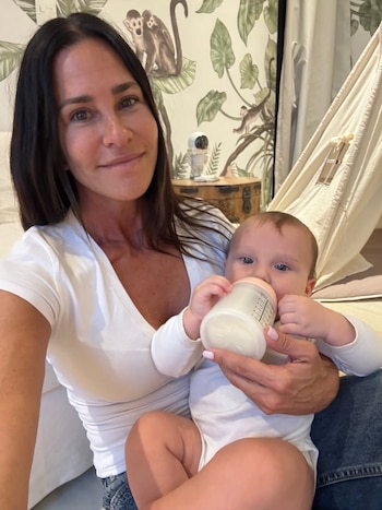 Carolina Baldini, de cabello oscuro, sonríe a cámara mientras sostiene a un bebé que bebe de un biberón, con un fondo de papel tapiz de monos y una tienda