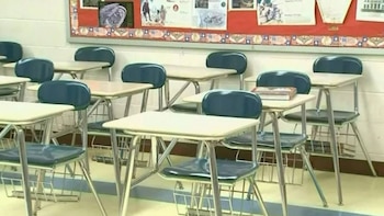 Un aula escolar vacía con pupitres y sillas azules con bases de metal organizados en filas ordenadas. Un tablón de anuncios con papeles se ve en la pared del fondo