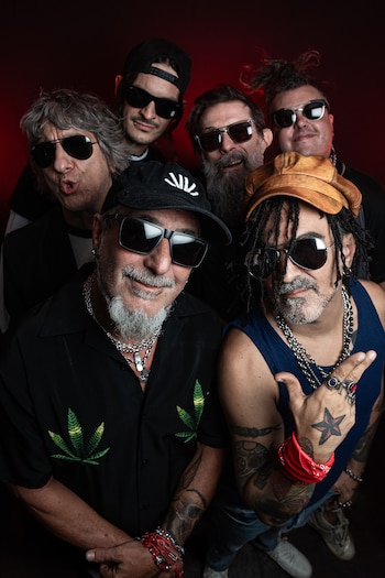 Seis hombres con gafas de sol, gorras y tatuajes miran a la cámara agrupados ante un fondo rojo