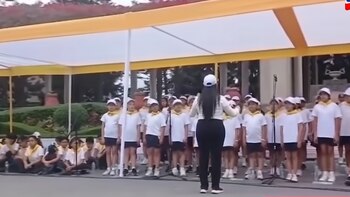 Una directora dirige a un grupo de niños con camisetas blancas y gorras alineados frente a micrófonos bajo un toldo amarillo y blanco