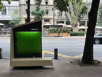 Un fotobiorreactor de color beige y negro con una pantalla grande que contiene un líquido verde brillante, ubicado en una acera urbana junto a árboles y edificios