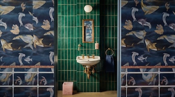 Un baño con papel tapiz de animales marinos, pared de azulejos verdes esmeralda, lavabo blanco con grifería dorada, espejo y toallero con toalla azul.