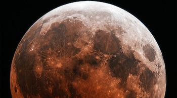 Una imagen macro de la Luna que presenta un eclipse lunar parcial profundo, con su superficie en tonos rojizos y grises, y una pequeña sección brillante.