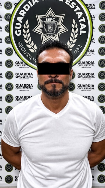 Hombre con cabello oscuro y barba, vistiendo camiseta blanca con cuello en V, posa frente a un fondo con logos de la Guardia Civil Estatal. Ojos cubiertos por barra negra