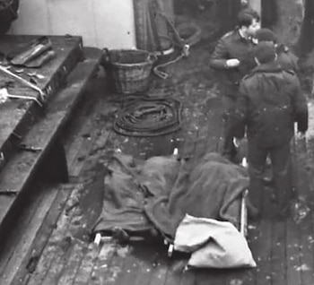 Tres hombres en uniforme permanecen en la cubierta de madera de una embarcación junto a camillas cubiertas con mantas