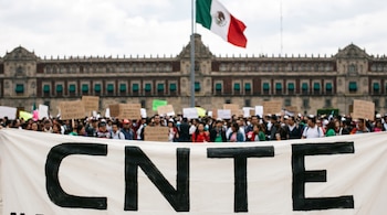 Primer plano de una pancarta blanca con "CNTE" en letras negras; detrás, una multitud frente al Palacio Nacional de México con la bandera nacional.
