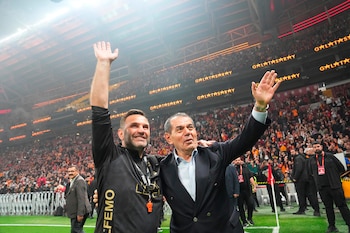 Dos hombres sonrientes saludan a la multitud en un estadio de fútbol del Galatasaray. Uno lleva un chándal negro y un silbato, el otro un traje oscuro