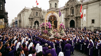 Una multitud vestida con túnicas moradas y velos blancos acompaña un anda procesional con flores frente a una iglesia de piedra.