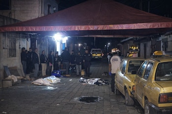 Vista nocturna de una calle adoquinada con personas, policías, personal de emergencia, dos vehículos amarillos y dos cuerpos cubiertos bajo una lona roja.