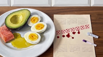 Un plato con medio aguacate, huevos cocidos, salmón y aceite junto a un calendario marcado con tinta roja, una lanceta médica y una tira reactiva.