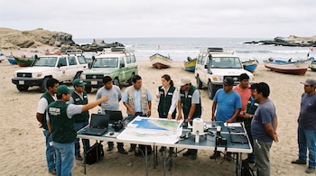Personas en una playa reunidas junto a una mesa con mapas, computadoras portátiles y cámaras. Hay vehículos todoterreno y botes de pesca en la arena.