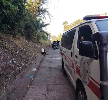 Una ambulancia blanca con franjas rojas estacionada en el carril de una carretera junto a vegetación y varias personas al fondo