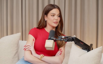 Khloé Kardashian, con cabello castaño y camiseta roja, sentada con los brazos cruzados frente a un micrófono de podcast que muestra su logo