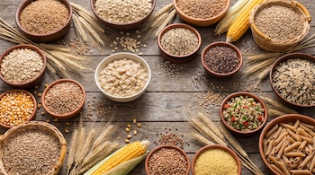 Múltiples cuencos y cestas con granos enteros como avena, trigo, quinoa, arroz integral, maíz y cebada. Espigas de trigo y mazorcas de maíz en mesa de madera.