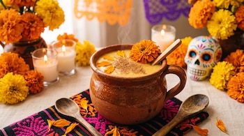 Taza de barro con atole amarillo y flores de cempasúchil, junto a velas encendidas, una calavera decorada y cucharas de madera sobre una mesa.