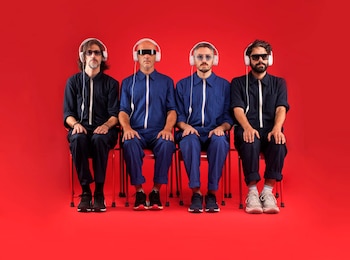Cuatro hombres sentados en sillas rojas. Visten overoles azules y negros, auriculares blancos y gafas. Fondo rojo liso