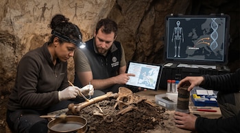 Personas examinan huesos con pinceles en una cueva, junto a una tableta digital, un cilindro de sedimento y una pantalla con mapas y esquemas.