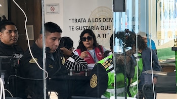 Interior de una comisaría con varias personas, incluidos policías uniformados y una mujer con un chaleco rojo del Ministerio de la Mujer, sentados tras un vidrio