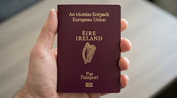 Primer plano de una mano sosteniendo un pasaporte irlandés de color granate. El pasaporte muestra el texto "European Union" e "Ireland" en dorado.