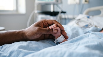 Una mano adulta sujeta la mano pequeña de un recién nacido con pulsera blanca sobre una sábana azul de hospital.