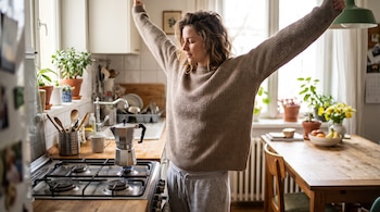 Una mujer en suéter y pants estira sus brazos en una cocina. Una cafetera moka está sobre la estufa con una llama. Se ven plantas, una mesa con frutas y flores.