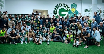 Gran grupo de hombres y algunos niños posan en uniforme de fútbol verde y blanco sobre césped artificial, con el logo del Goiás Esporte Clube al fondo
