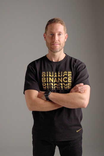 Hombre con barba rubia, camiseta negra con el logo de Binance en dorado y la palabra BINANCE repetida, reloj digital negro en la muñeca izquierda, brazos cruzados