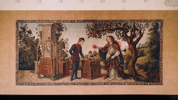Tapiz intrincado que muestra a un botones uniformado junto a maletas y una mujer con cabello largo ofreciéndole una granada bajo un árbol de manzanas