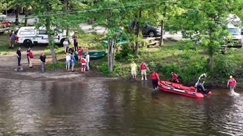 Vista aérea de personas en la orilla y en el agua, un bote inflable rojo en el río y botes verdes en la hierba, rodeados de árboles y vehículos