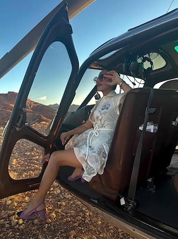 Mujer con gafas de sol y vestido blanco floral sentada en el asiento de un helicóptero con la puerta abierta, viendo un paisaje desértico montañoso