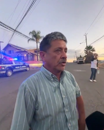 Hombre de mediana edad con camisa a rayas, una patrulla con luces encendidas, una calle, casas, postes, cables y palmeras