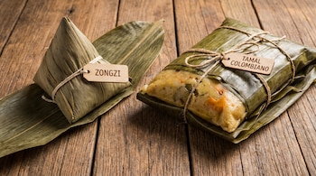 Vista superior de un zongzi en forma triangular a la izquierda y un tamal colombiano rectangular a la derecha, ambos envueltos en hojas y sobre madera.