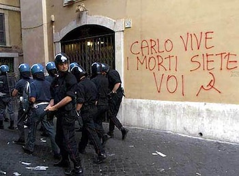 Grupo de policías con uniformes negros y cascos azules camina por una calle. Un muro de edificio color arena presenta un grafiti rojo