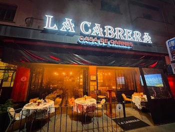 Fachada de restaurante La Cabrera Casa de Carnes con letrero luminoso, mesas y sillas dispuestas en terraza y salón, y luces encendidas por la noche