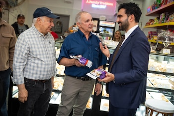 Tres hombres conversan junto a vitrinas con repostería en un comercio; dos sostienen pequeños paquetes embalados