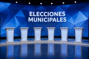 Seis atriles blancos con micrófonos frente a una pantalla azul con el texto Elecciones Municipales.