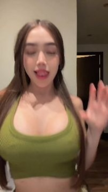 Mujer joven, cabello castaño en coletas, top verde, ojos cerrados, lengua ligeramente afuera, una mano levantada, fondo claro