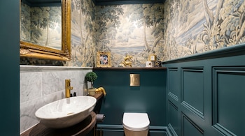 Baño de diseño elegante con tapiz de paisaje boscoso y río en paredes superiores, y revestimiento teal oscuro abajo. Incluye lavabo de mármol blanco, grifo dorado, inodoro y espejo.